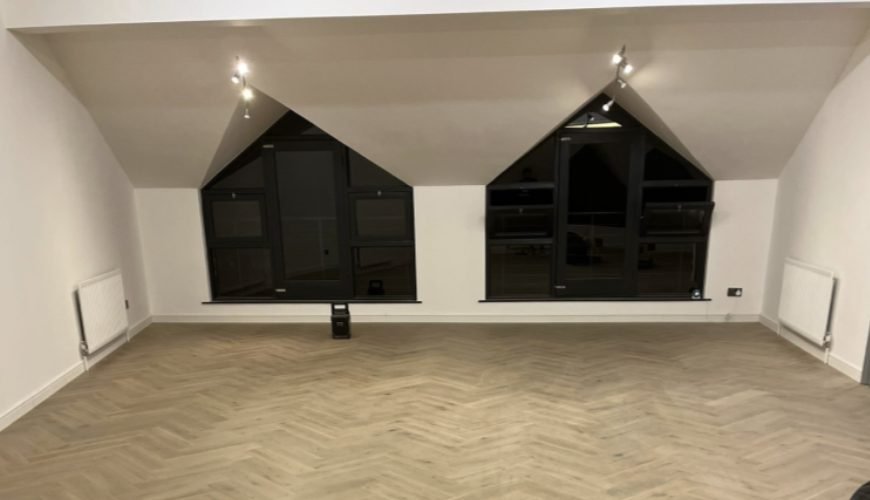 Large open loft room with two apex gable windows, herringbone flooring, track lighting and radiators
